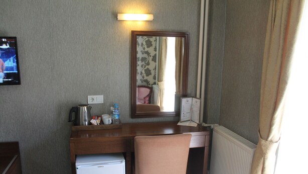 A hotel room with a wooden desk, a small refrigerator, a chair, a mirror, and a window with curtains. - Rest Hotel (Ankara)