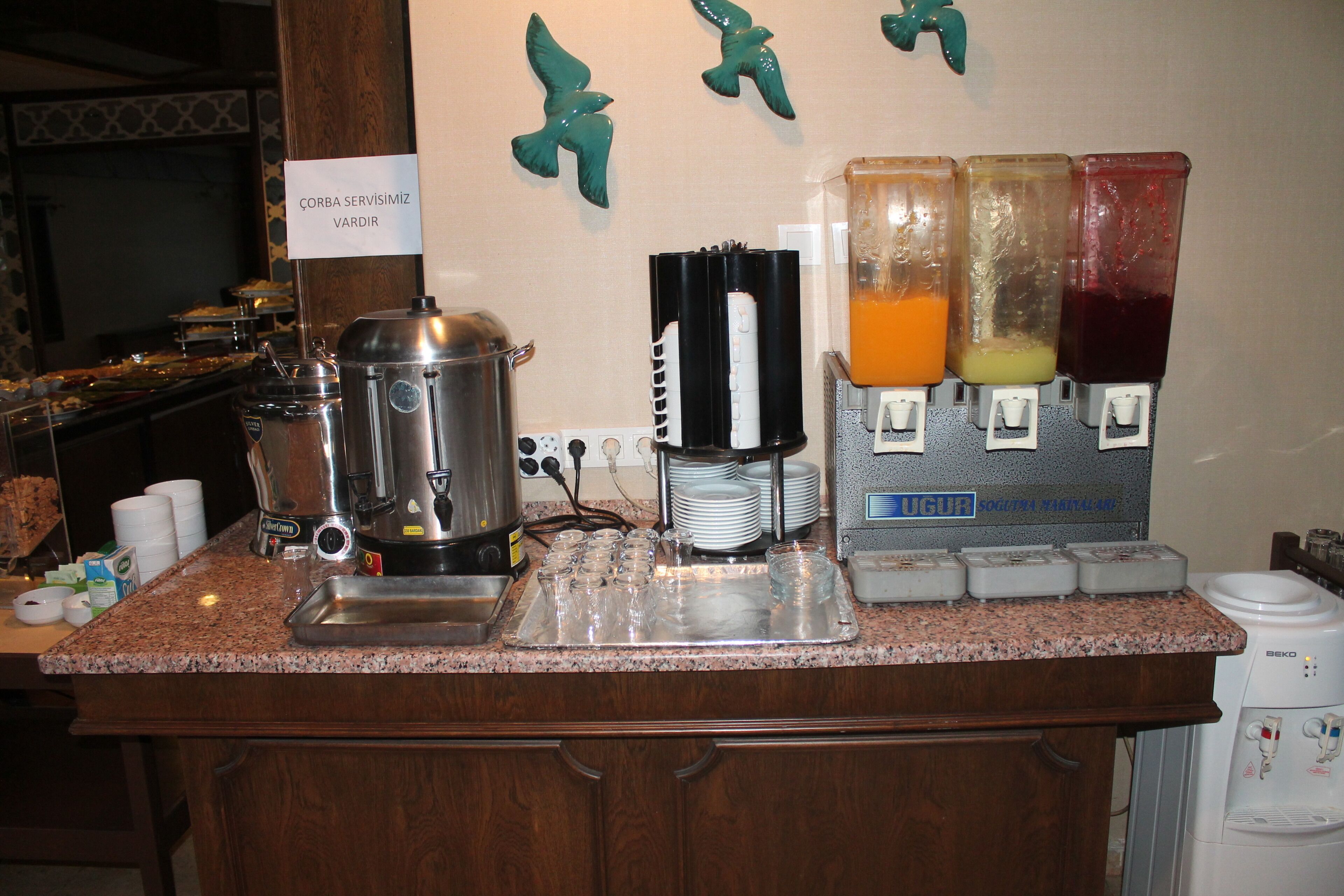 A buffet setup with a coffee maker, juice dispensers, and a sign indicating 'CORBA SERVISIMIZ VARDIR'.