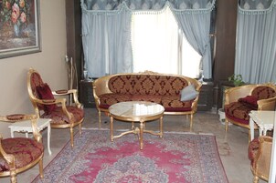 Una habitación con un sofá rojo estampado, sillas doradas y rojas, una mesa de centro blanca y un ventanal con cortinas azul claro.