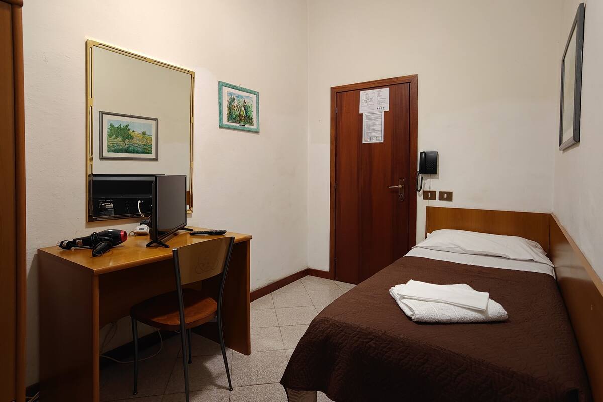 A hotel room with a wooden desk, a computer, a chair, a bed with a brown bedspread, and a door with a notice on it.