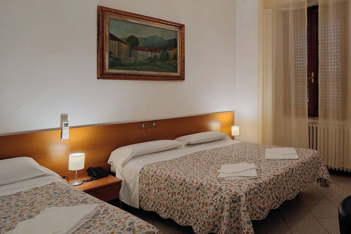 A hotel room with two beds, a painting on the wall, a bedside lamp, and a window with blinds.