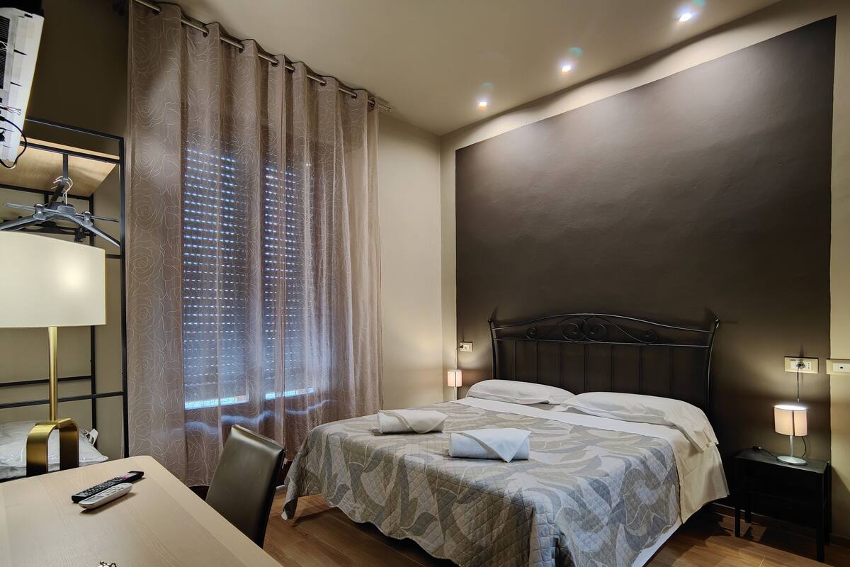 A hotel room with a bed, bedside tables, a desk with a lamp, and a window with curtains.