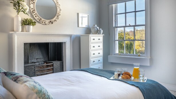 Une chambre avec une cheminée, une commode, une fenêtre donnant sur un beau paysage et un plateau de petit-déjeuner posé sur le lit.