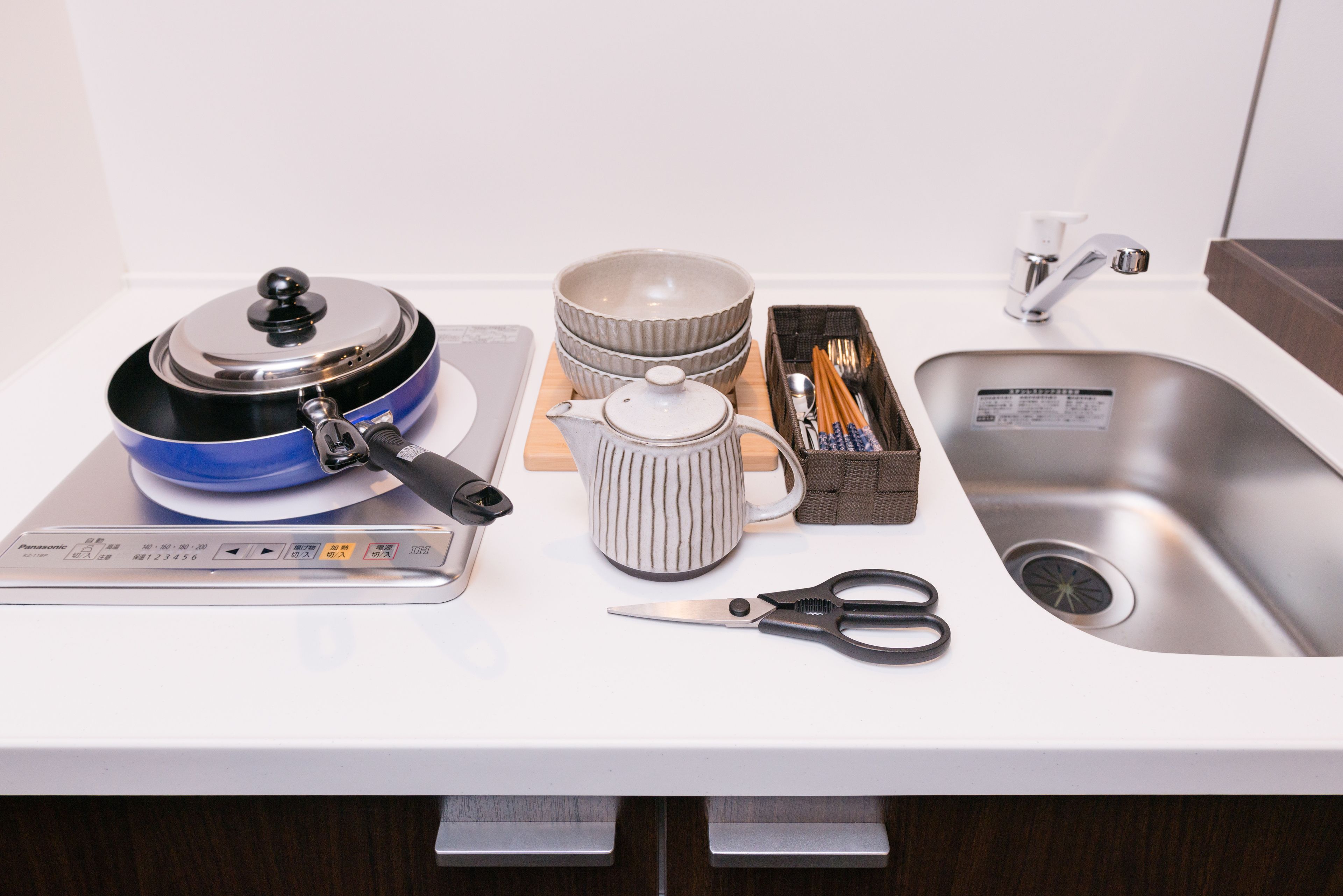 A kitchen counter with a rice cooker, bowls, a teapot, a box of chopsticks, and a pair of scissors.