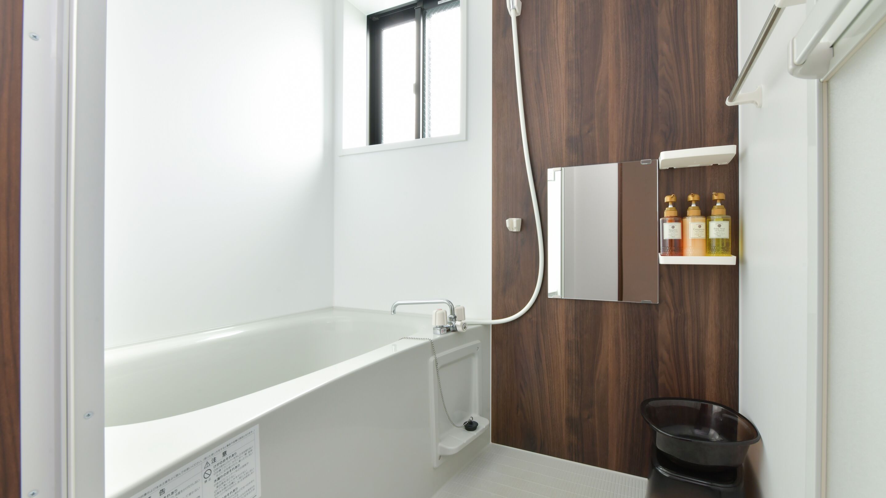 Een moderne badkamer met een vrijstaand bad, een wandpaneel van hout, een inbouwkastje met toiletartikelen en een zwarte prullenbak.