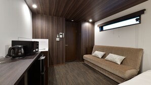 A compact hotel room with a wooden ceiling, a sofa with floral cushions, a desk with a kettle, and a window with blinds. - Villa Front Kyoto Seimei (Kyoto)