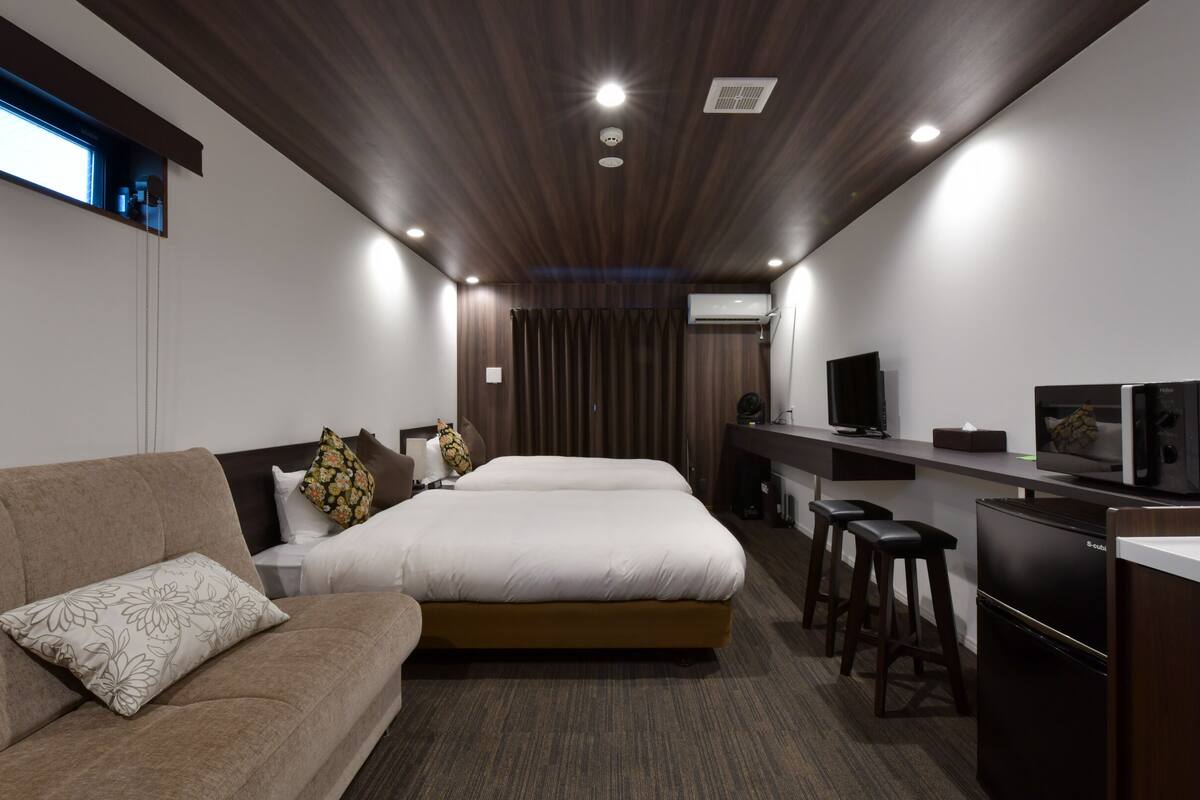 A modern hotel room with a large bed, a sofa, a desk with a computer and a TV, and a kitchenette.