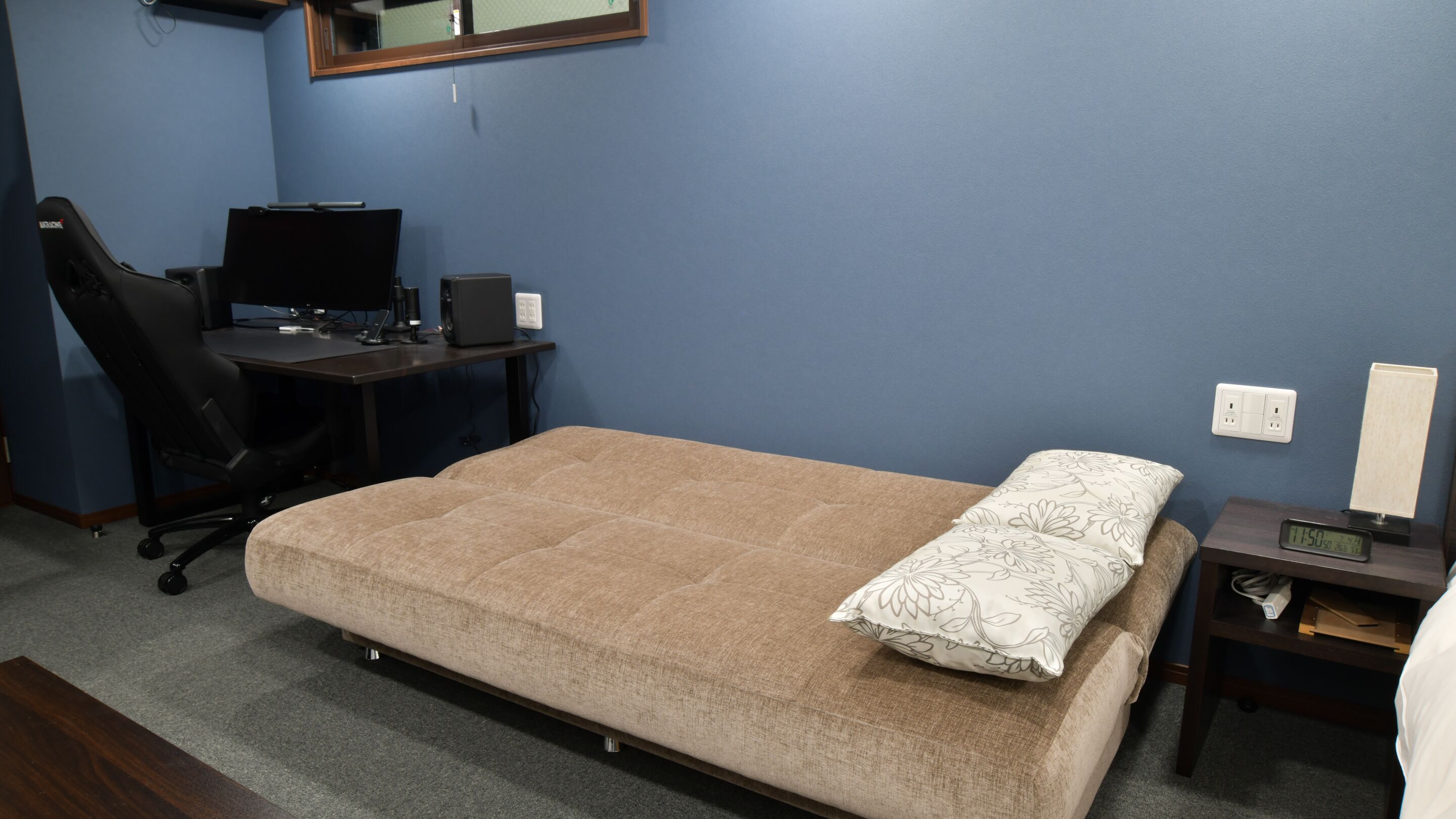 A bedroom with a bed, a desk with a computer, a chair, and a nightstand with a clock.