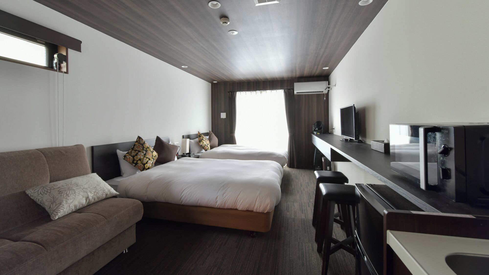 A modern hotel room with a sofa, two beds, a desk with a computer, and a TV.
