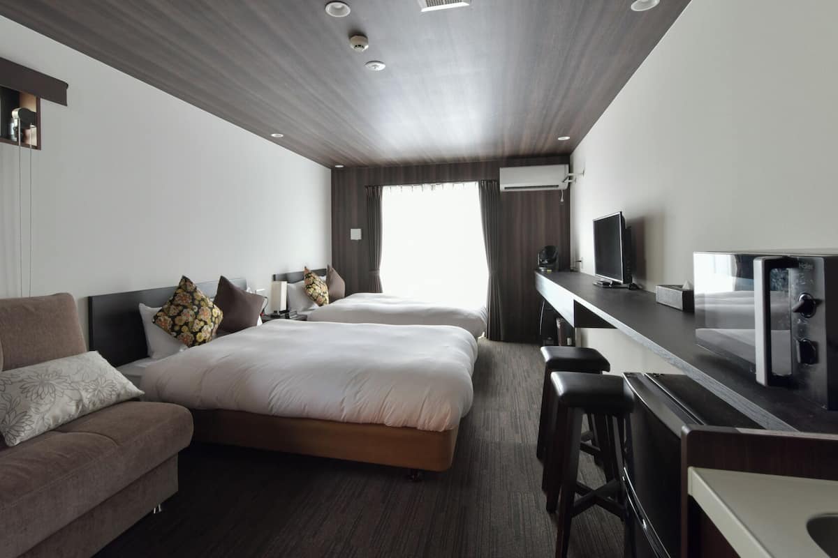 A modern hotel room with a sofa, two beds, a desk with a computer, and a TV.