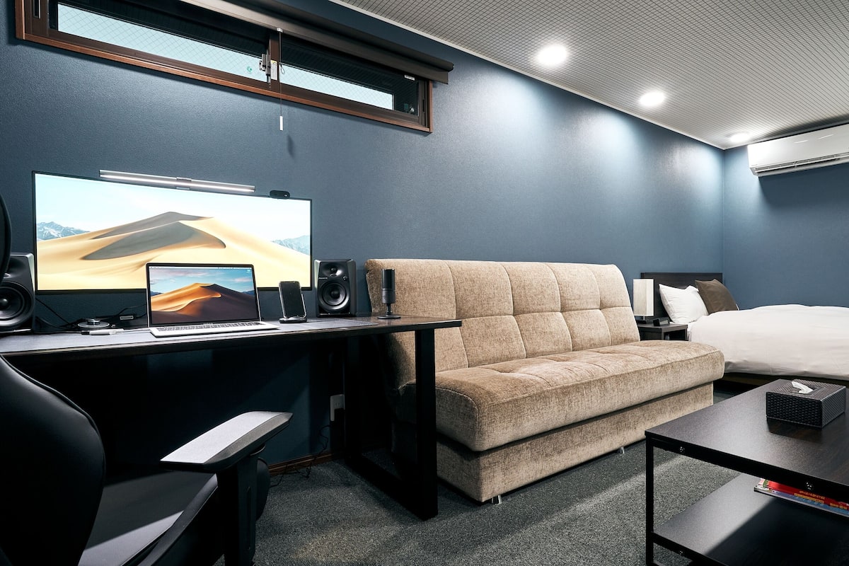 A modern hotel room with a desk, laptop, and smartphone, a sofa, a bed, and a small table with a remote control.