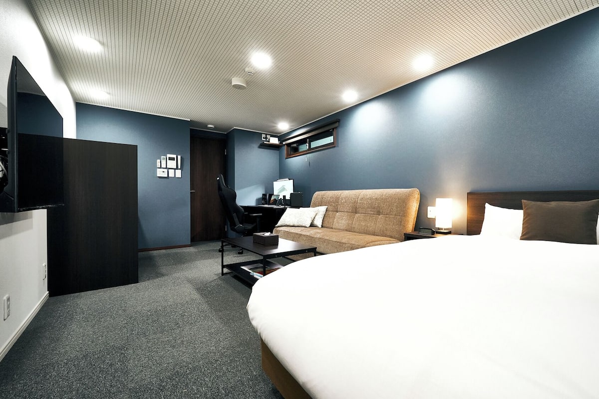 A hotel room with a large bed, a sofa, a desk with a computer, and a TV mounted on the wall.