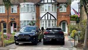 A residential street with two cars parked in front of a house with a white facade and black accents.