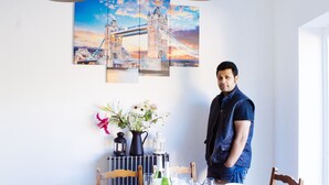 A dining area with a table set for two, a vase of flowers, and a wall art piece of a bridge.