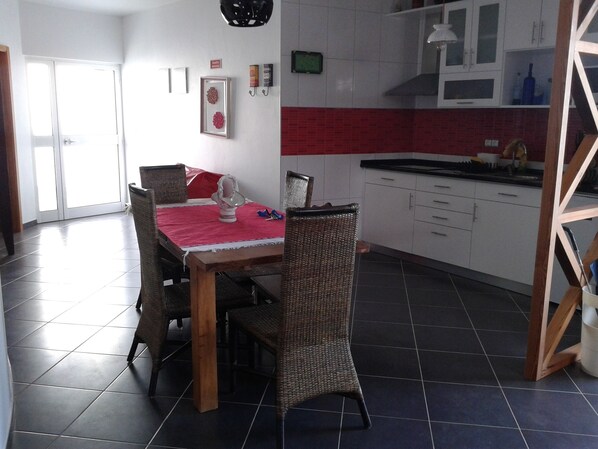 A dining area with a table set for four, surrounded by wicker chairs. A red tablecloth covers the table, and there are two framed decorative items on the wall.