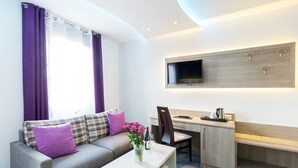 A modern hotel room with a grey sofa, purple cushions, a white coffee table, a desk with a chair, and a mounted TV. - Bavaria Hotel Superior (Stuttgart)