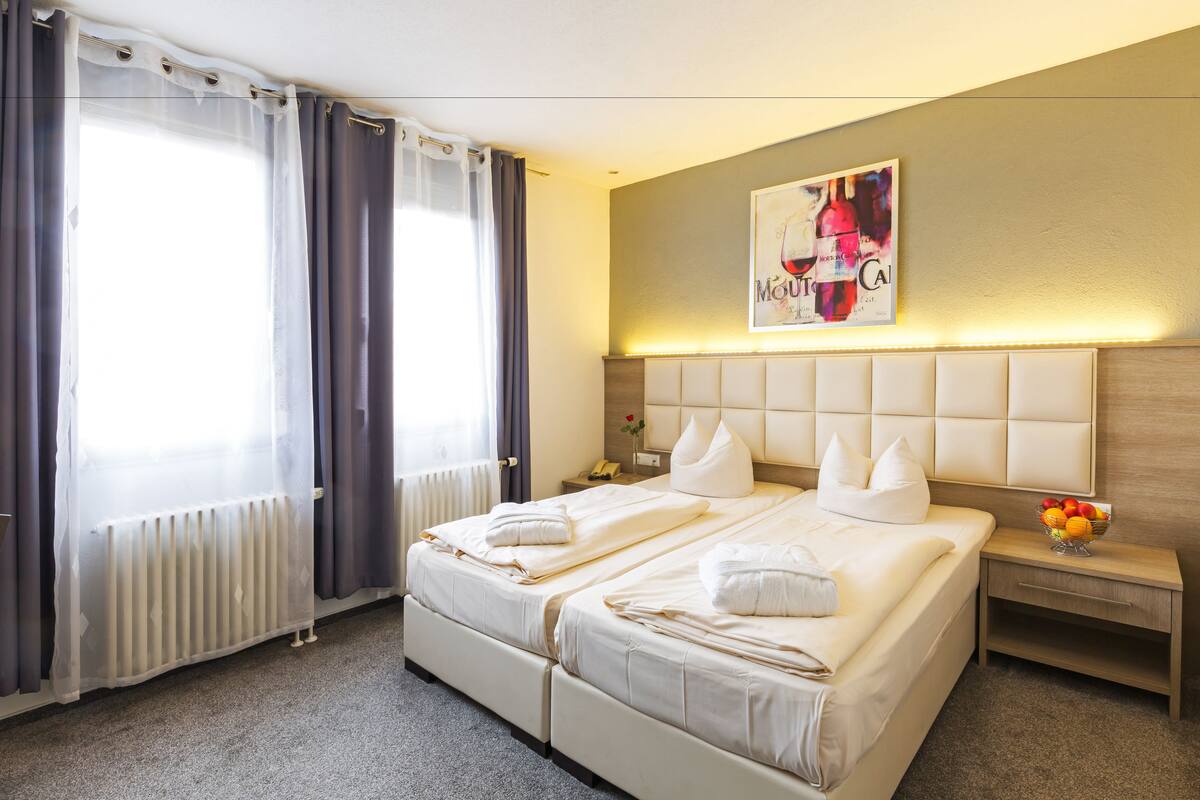 A hotel room with two beds, a bedside table with a fruit bowl, a wall-mounted artwork, and a radiator.