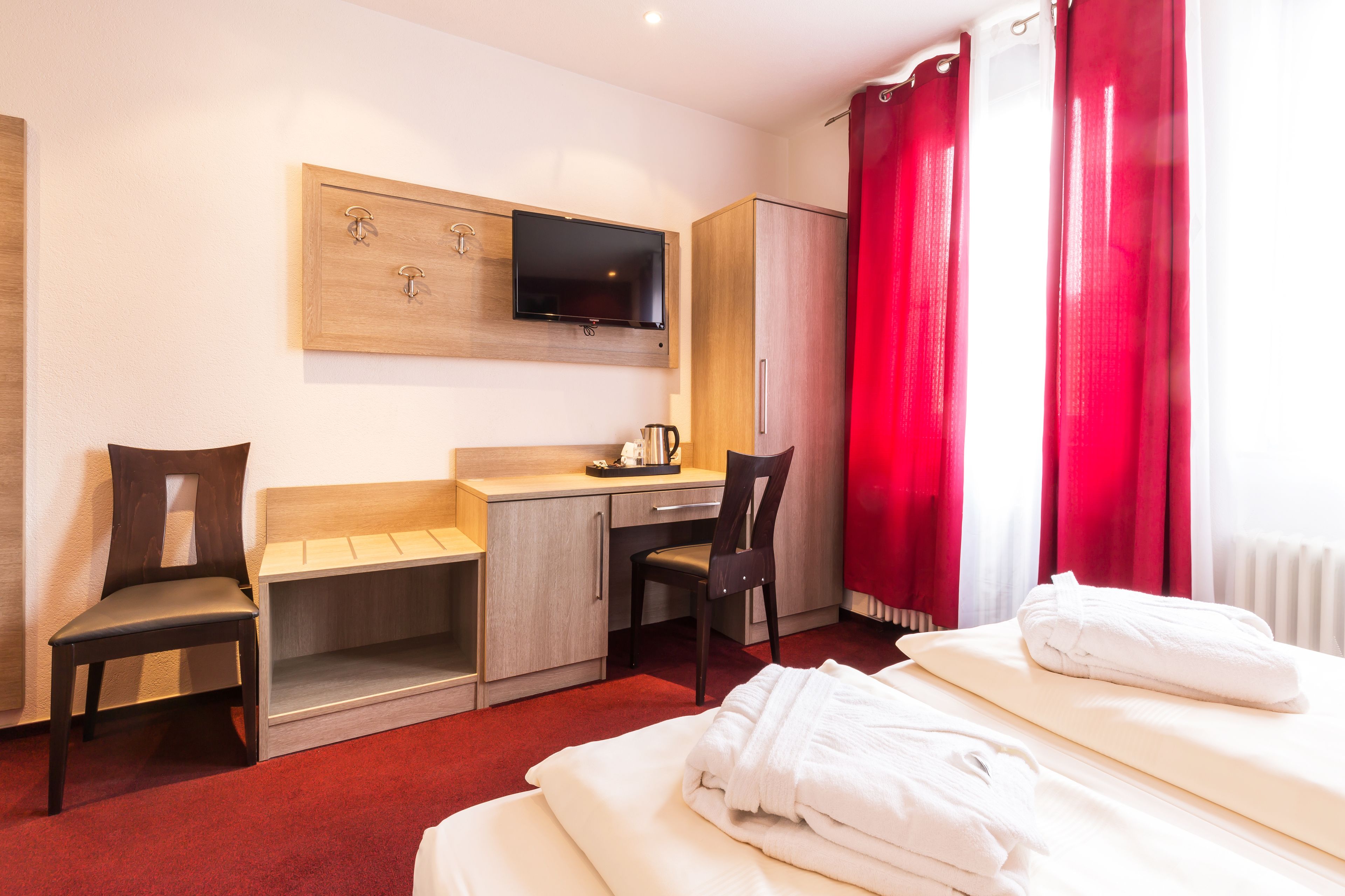 A hotel room with a bed, a desk, a chair, a TV mounted on the wall, and red curtains.