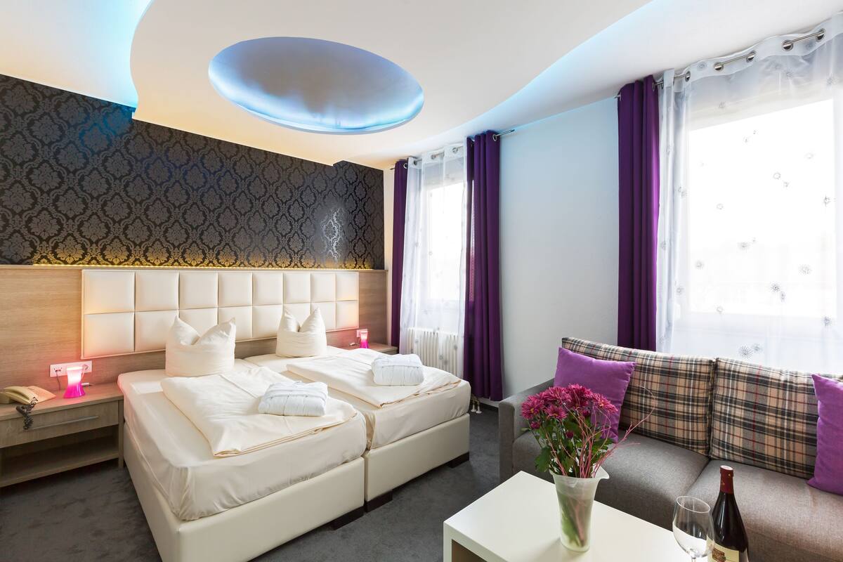A hotel room with a double bed, a sofa, a bedside table with a phone and lamp, and a window with curtains.