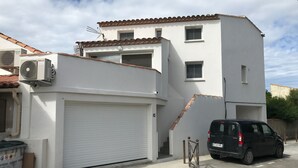A two-story building with a white facade, a garage door, and a parked black van. - Luxury seafront apartment 4**** First floor (Saintes-Maries-De-La-Mer)