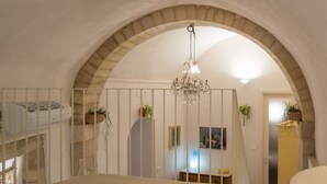 Un dormitorio con una cama grande, techo de piedra abovedado, una araña y vista a una cocina por una ventana con barrotes.