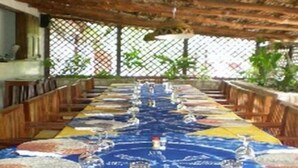 A long dining table with chairs, covered by a blue tablecloth with a pattern, in a covered outdoor area with a wooden ceiling and a fence in the background. - Le Calao du Lac Rose (Niaga)