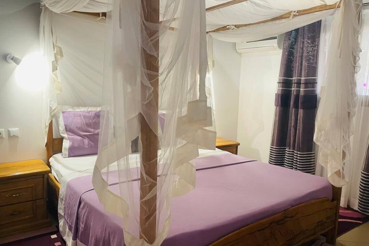 Ein Schlafzimmer mit einem Himmelsbett, einem Nachttisch aus Holz, einem Beistelltisch und einem Fenster mit Vorhängen.