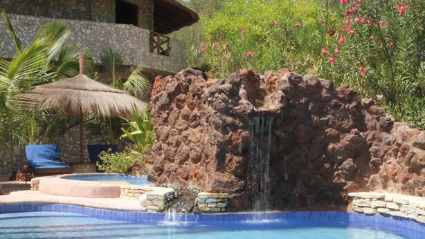 A pool with a waterfall feature, surrounded by a stone wall and lush greenery. - Le Calao du Lac Rose (Niaga)
