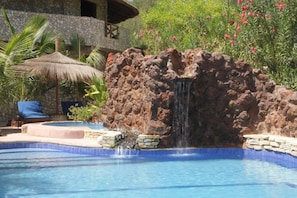 A pool with a waterfall feature, surrounded by a stone wall and lush greenery. - Le Calao du Lac Rose (Niaga)