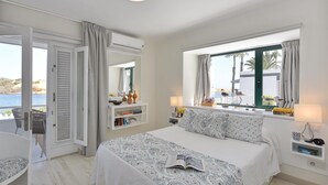 Un dormitorio con cama, mesita de noche, cortinas en la ventana y puerta que da a un balcón, con vista al mar.