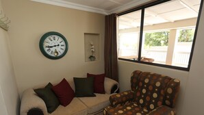 Un salon avec un canapé beige, un fauteuil à motifs, une horloge murale et une fenêtre donnant sur un balcon.