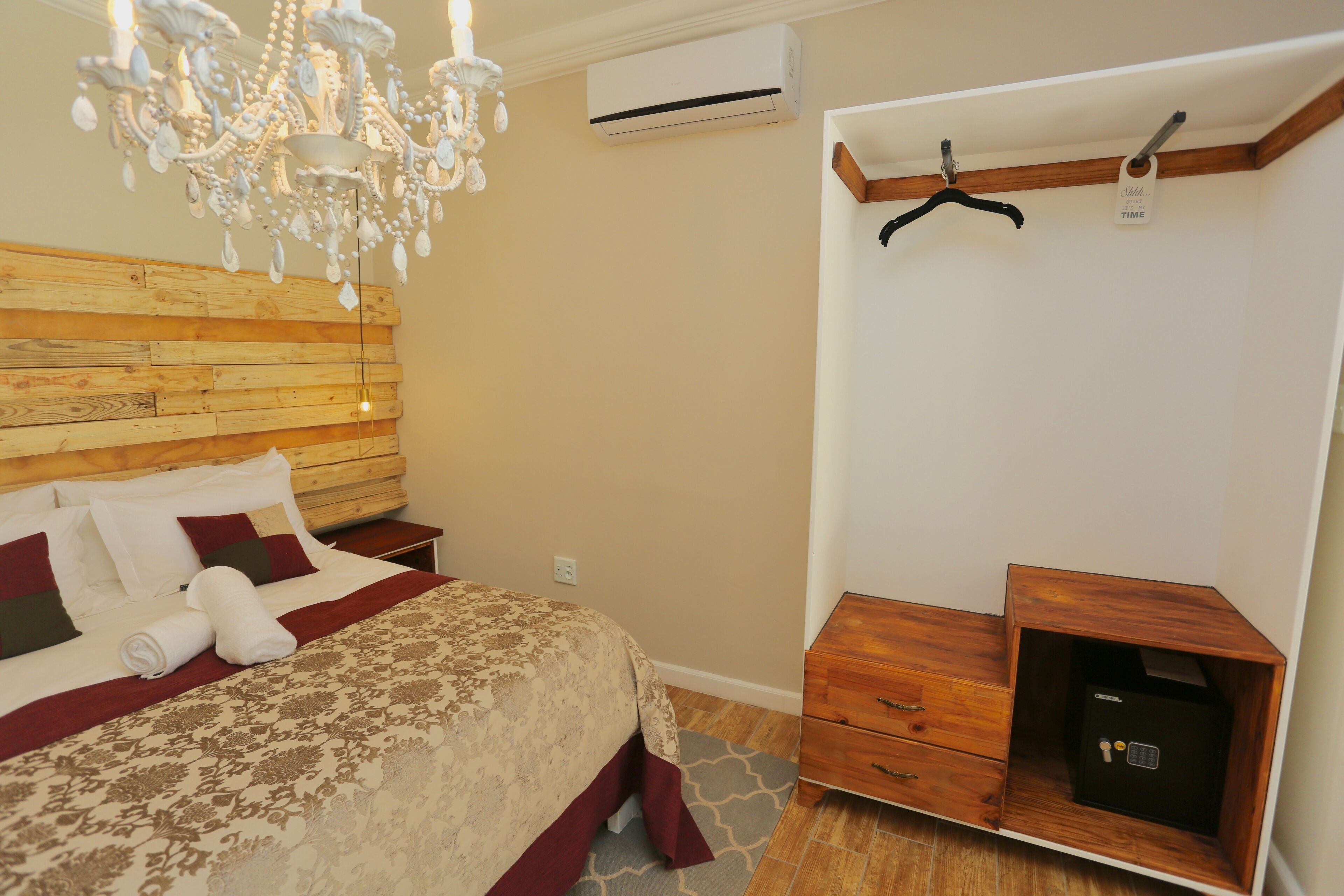 A bedroom with a wooden headboard, a chandelier, a bed with a patterned bedspread, a nightstand, a dresser with drawers, and a safe.