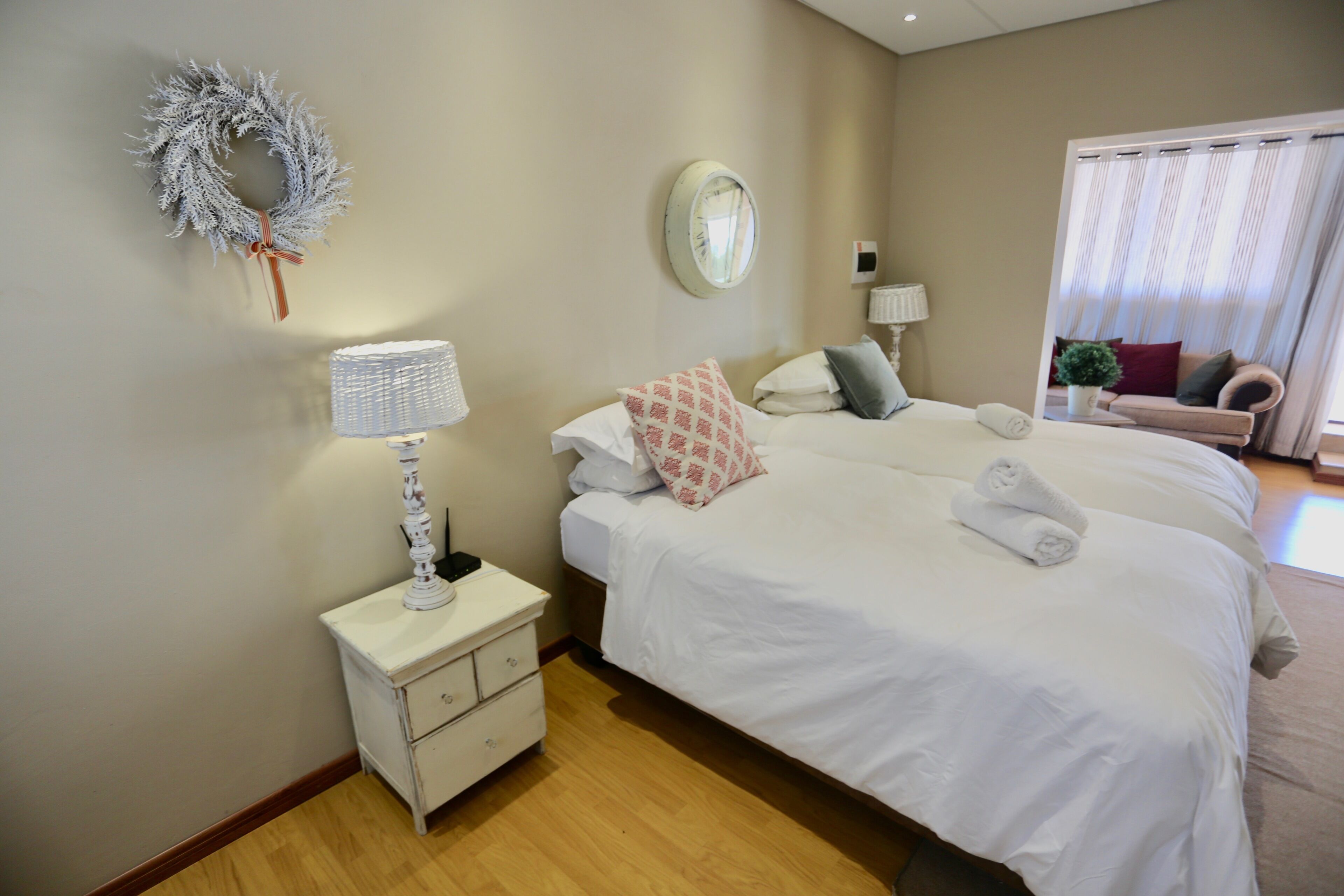 A hotel room with two beds, a nightstand with a lamp, a mirror, a wreath on the wall, and a window with curtains.