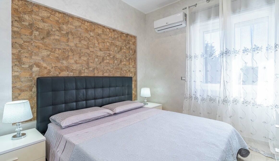 A bedroom with a stone accent wall, a bed with a black headboard, a nightstand with a lamp, and a window with sheer curtains.