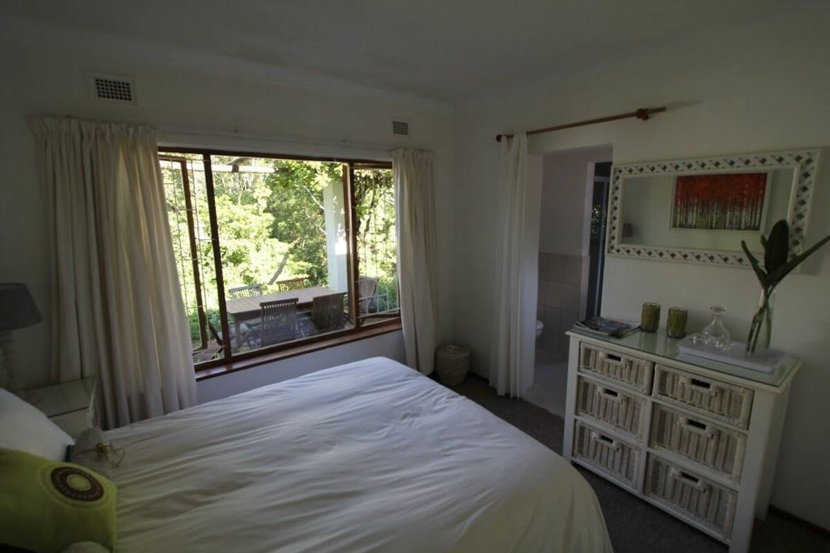 Un dormitorio con una cama grande, una ventana con cortinas, una mesita de noche con espejo y un armario pequeño.