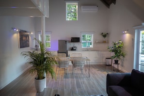 Una cocina moderna con armarios blancos, un comedor con mesa y sillas, y una sala de estar con sofá. Hay plantas en macetas y un televisor en la cocina.