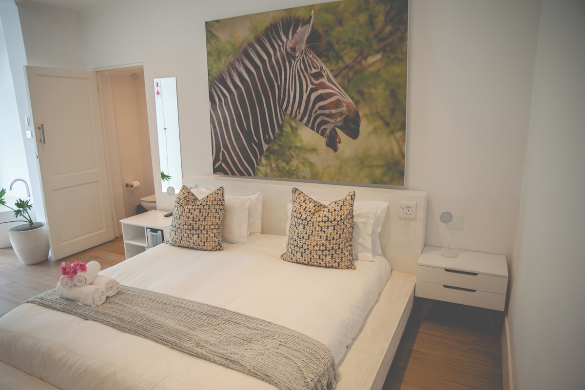 A hotel room with a large bed, two pillows, a bedside table, a nightstand, a wall-mounted painting of a zebra, a potted plant, and a towel on the bed.