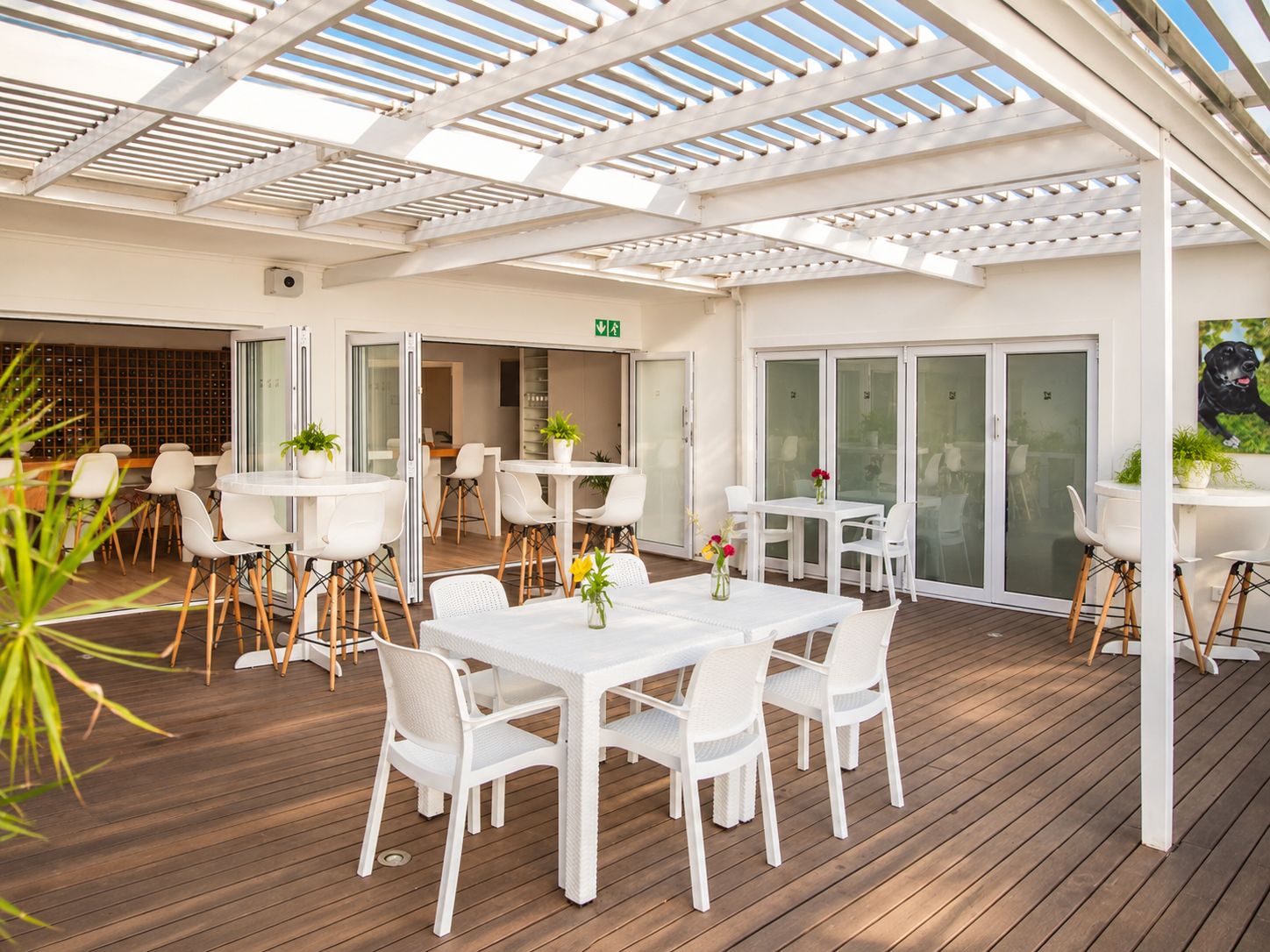 An outdoor patio area with white tables and chairs, a wooden deck, and a covered seating area with a white pergola.