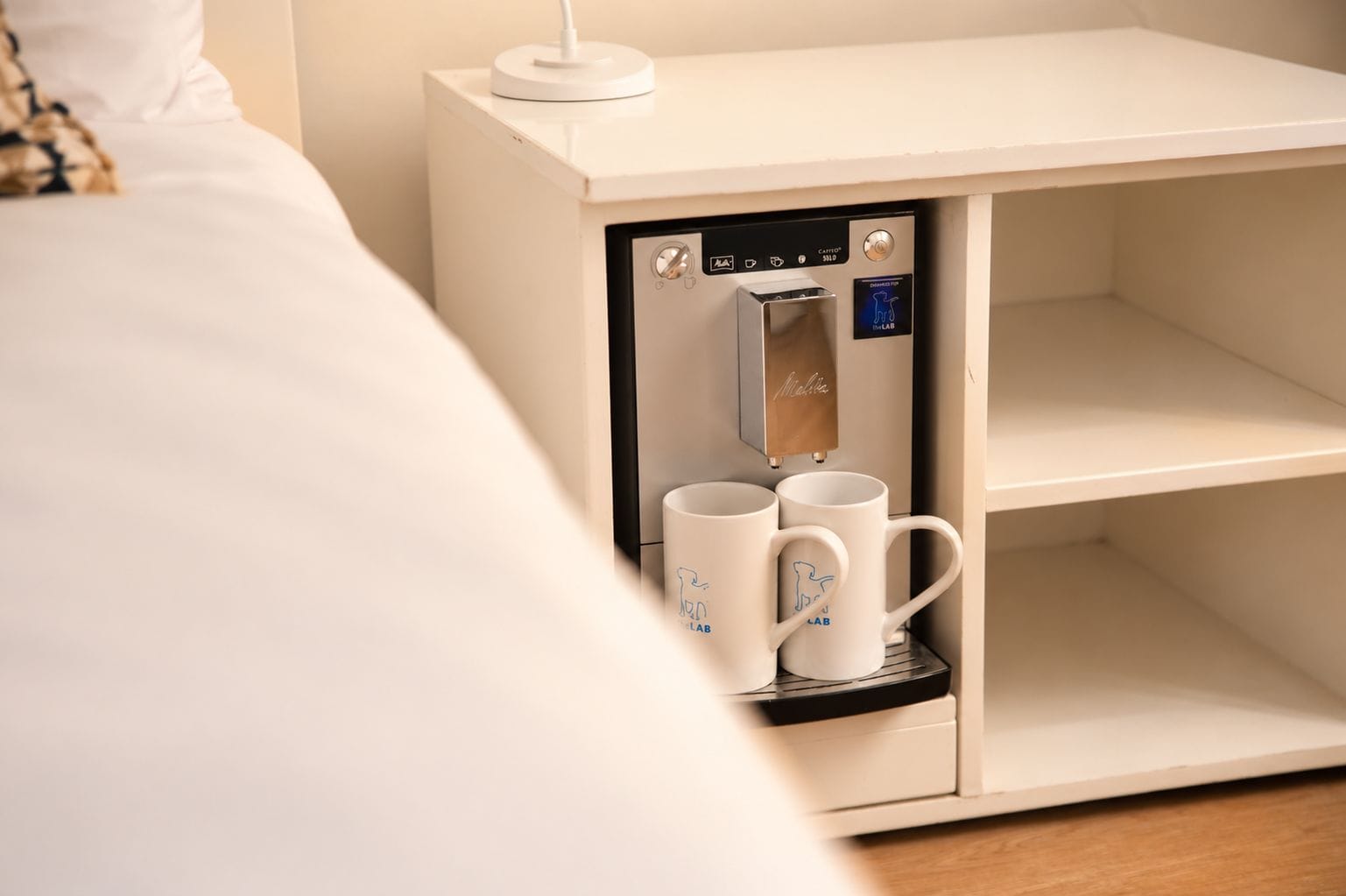 Une machine à café compacte dotée d’un écran numérique, deux tasses blanches ornées d’un motif de dinosaure et une table de chevet blanche.