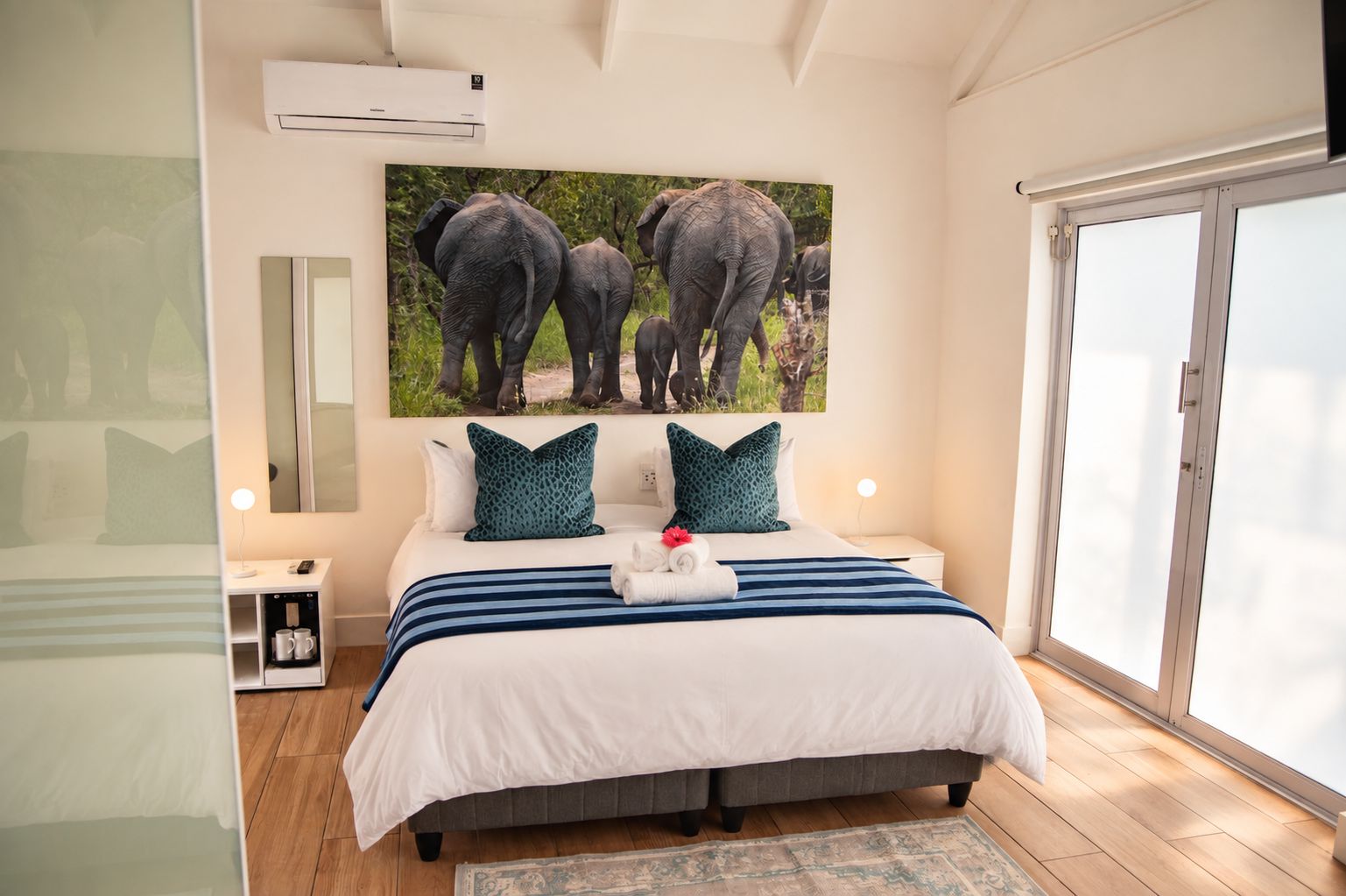 Una camera da letto con un letto grande, un comodino, una abat-jour, un condizionatore d'aria, una finestra e un quadro appeso al muro raffigurante degli elefanti.