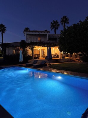 Una piscina con superficie de agua azul cristalina, rodeada por una terraza con mobiliario exterior y un edificio con entrada iluminada.