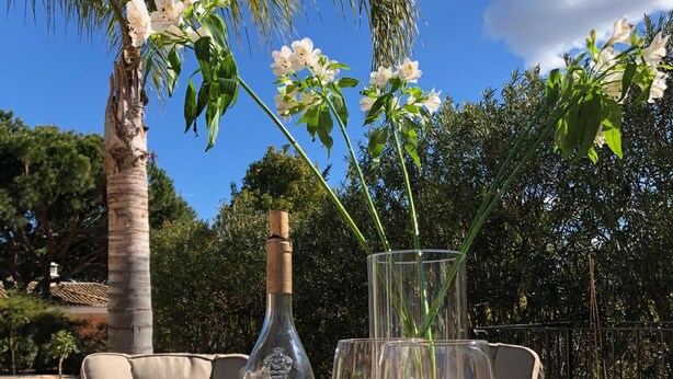 A wicker table with a bottle and two glasses of white wine, set outdoors with a palm tree and flowers in the background. - 4 Bedroom Deluxe Villa, Private Pool, Mature Grounds, Hot Tub*, Putting Green + (Quarteira)