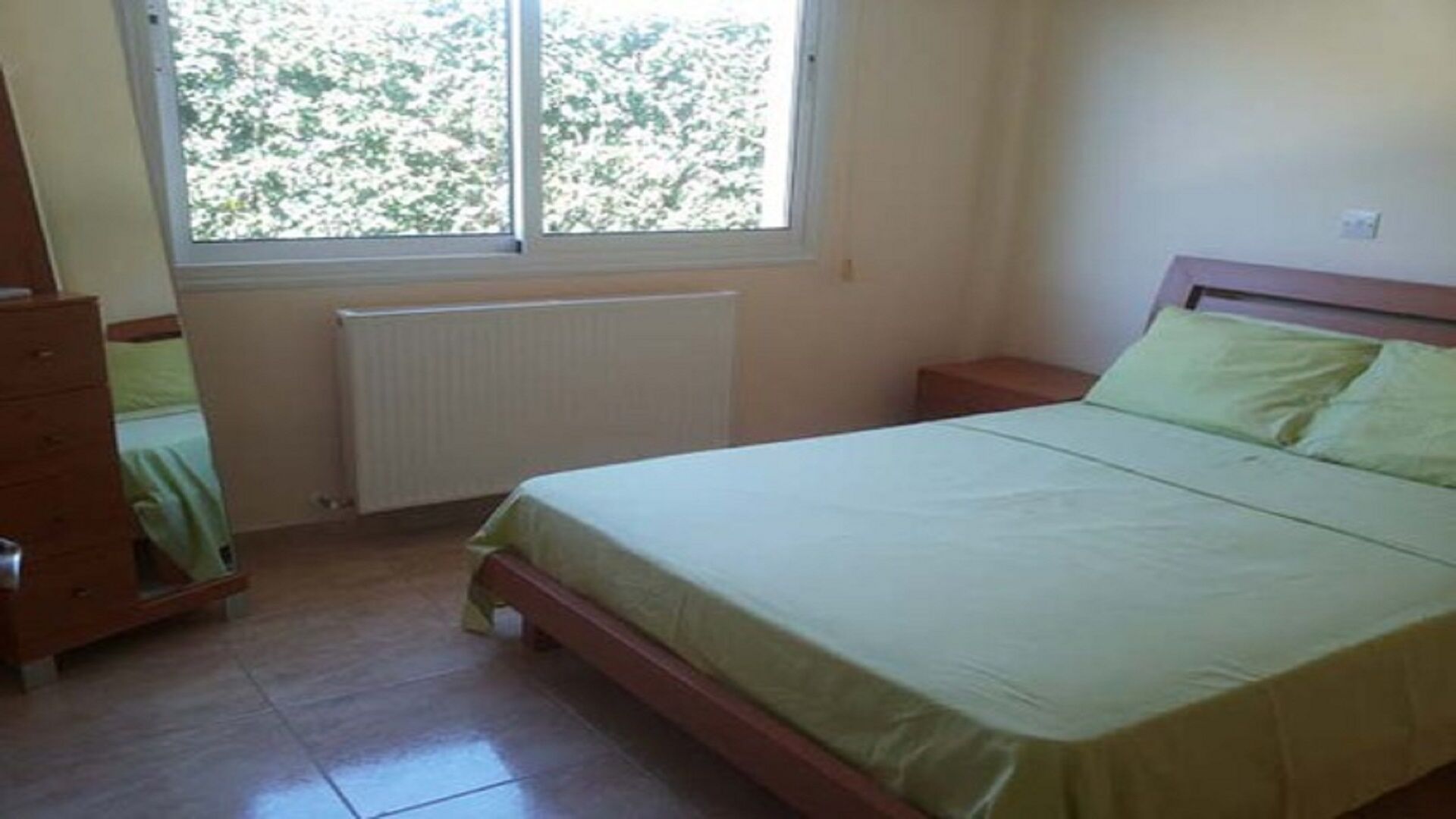 Een slaapkamer met een bed, een dressoir, een raam en een radiator.