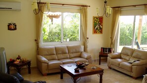 Ein Wohnzimmer mit beiger Couch, Couchtisch und Fernseher. Zwei Fenster mit Vorhängen, ein Bild an der Wand und ein Deckenventilator.