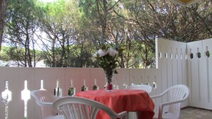 A covered patio with a table set for two, surrounded by white chairs, a red tablecloth, and a vase with flowers.