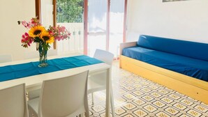 A room with a dining table, chairs, a sofa, a vase of flowers, and a view of greenery through the window.