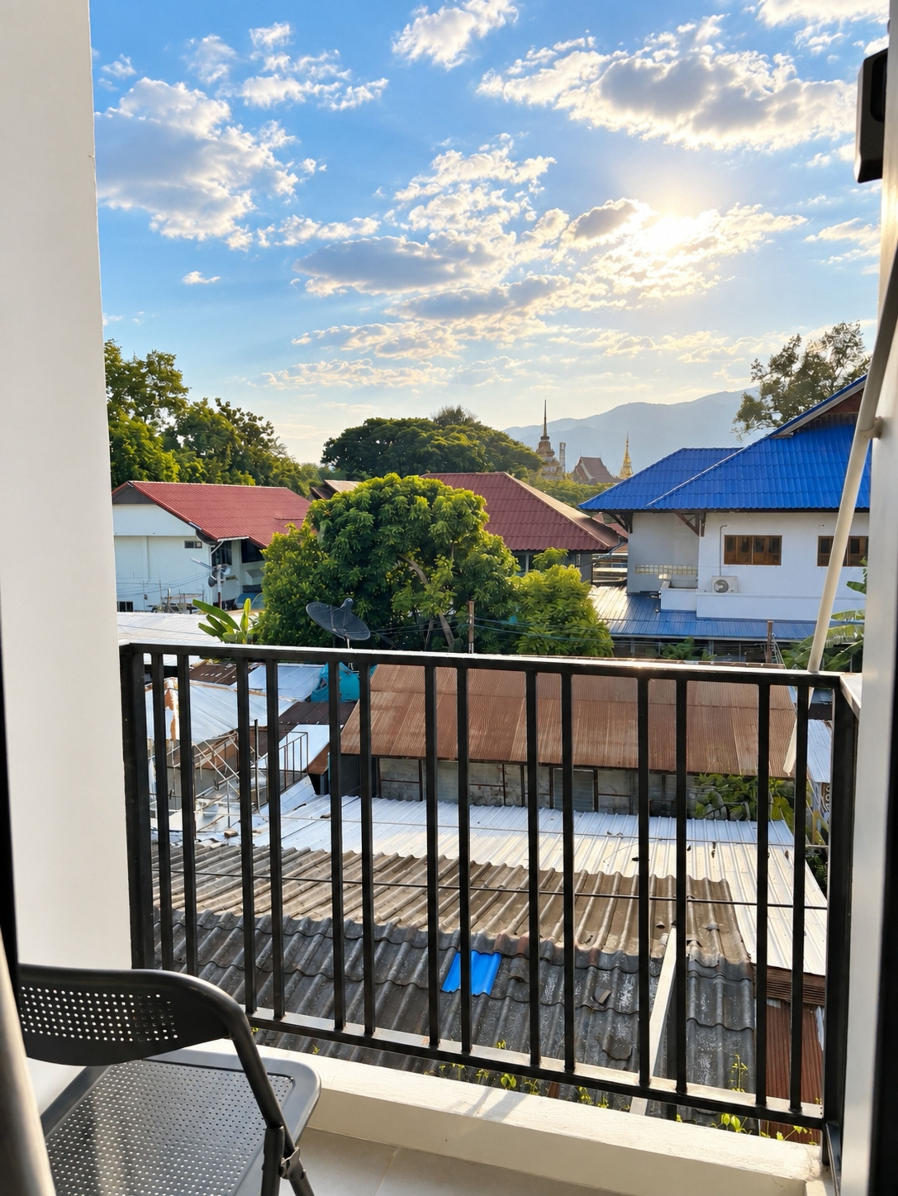Ein Balkon mit Metallgeländer, einem Stuhl und Blick auf Gebäude mit roten und blauen Dächern, einen Baum und einen klaren Himmel.
