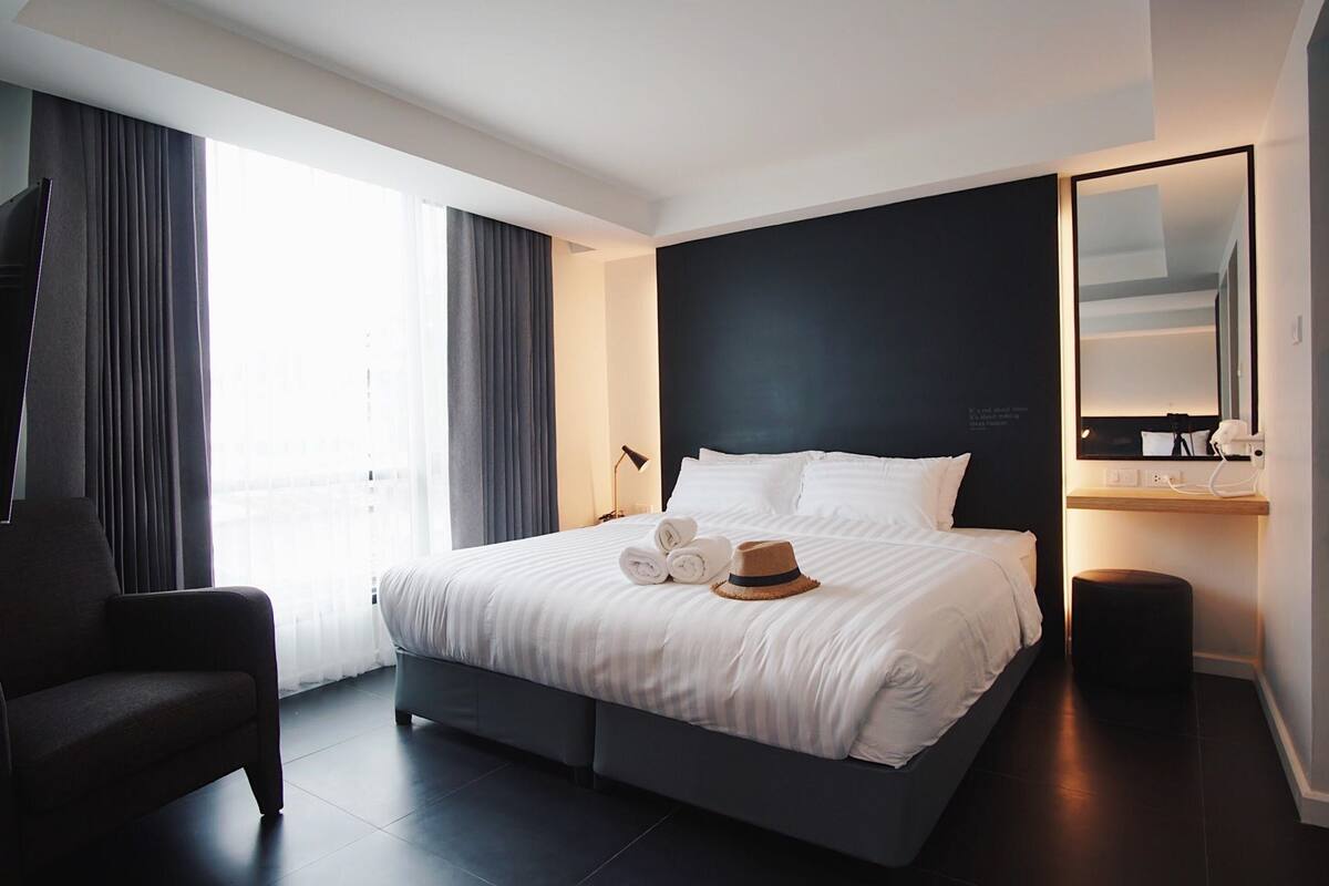 A modern hotel room with a large bed, a sitting area, a flat-screen TV, and a large window with curtains.
