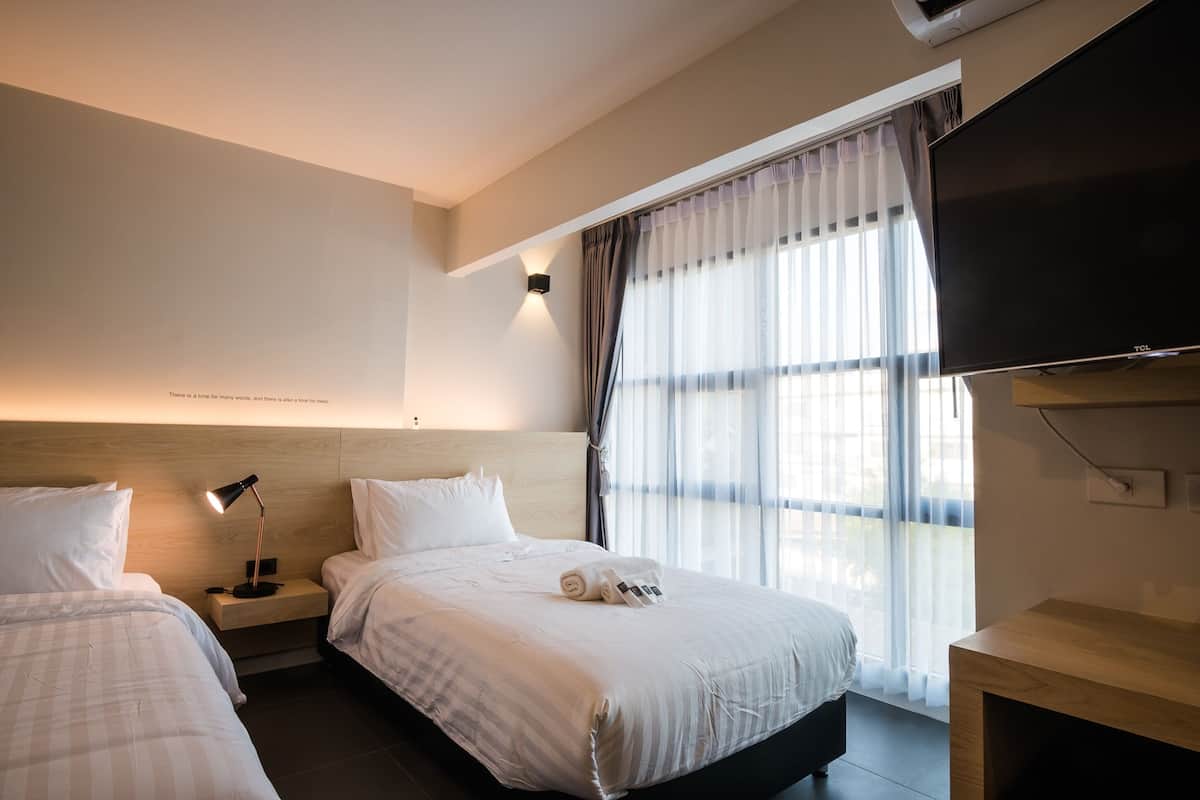 A hotel room with two beds, a flat-screen TV, a bedside table with a lamp, and a large window with sheer curtains.
