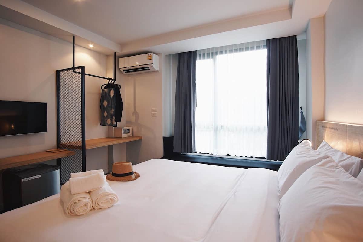 A hotel room with a neatly made bed, a TV mounted on the wall, an air conditioning unit, and a large window with curtains.